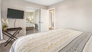 Een slaapkamer met een bed, een aan de muur bevestigde televisie, een klein tafeltje met een plant en een deur naar een andere kamer.