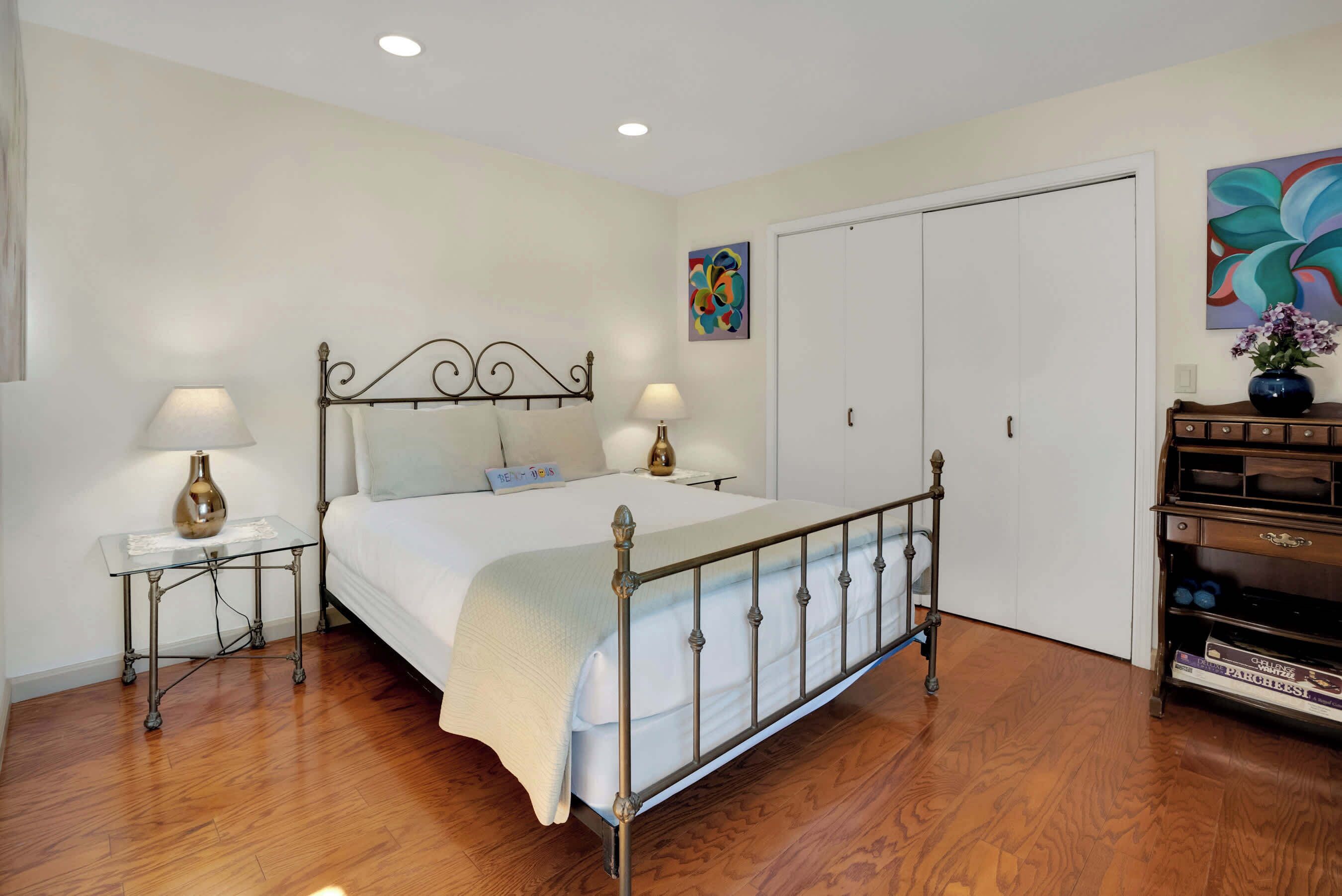 Un dormitorio con una cama de estructura metálica, dos mesitas de noche con lámparas, una puerta blanca y una pared con un cuadro abstracto colorido.