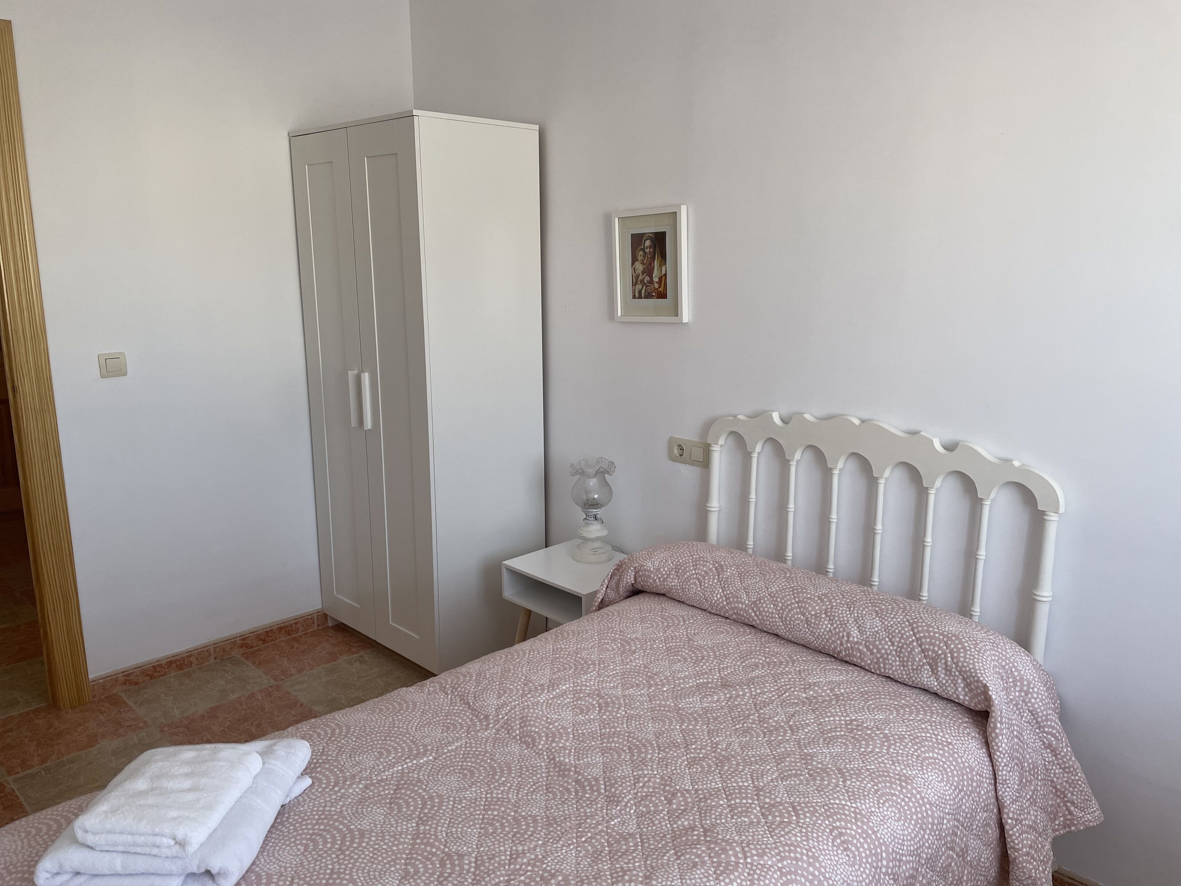 Un dormitorio con cama, un armario blanco, una mesita de noche con lámpara y un cuadro en la pared.
