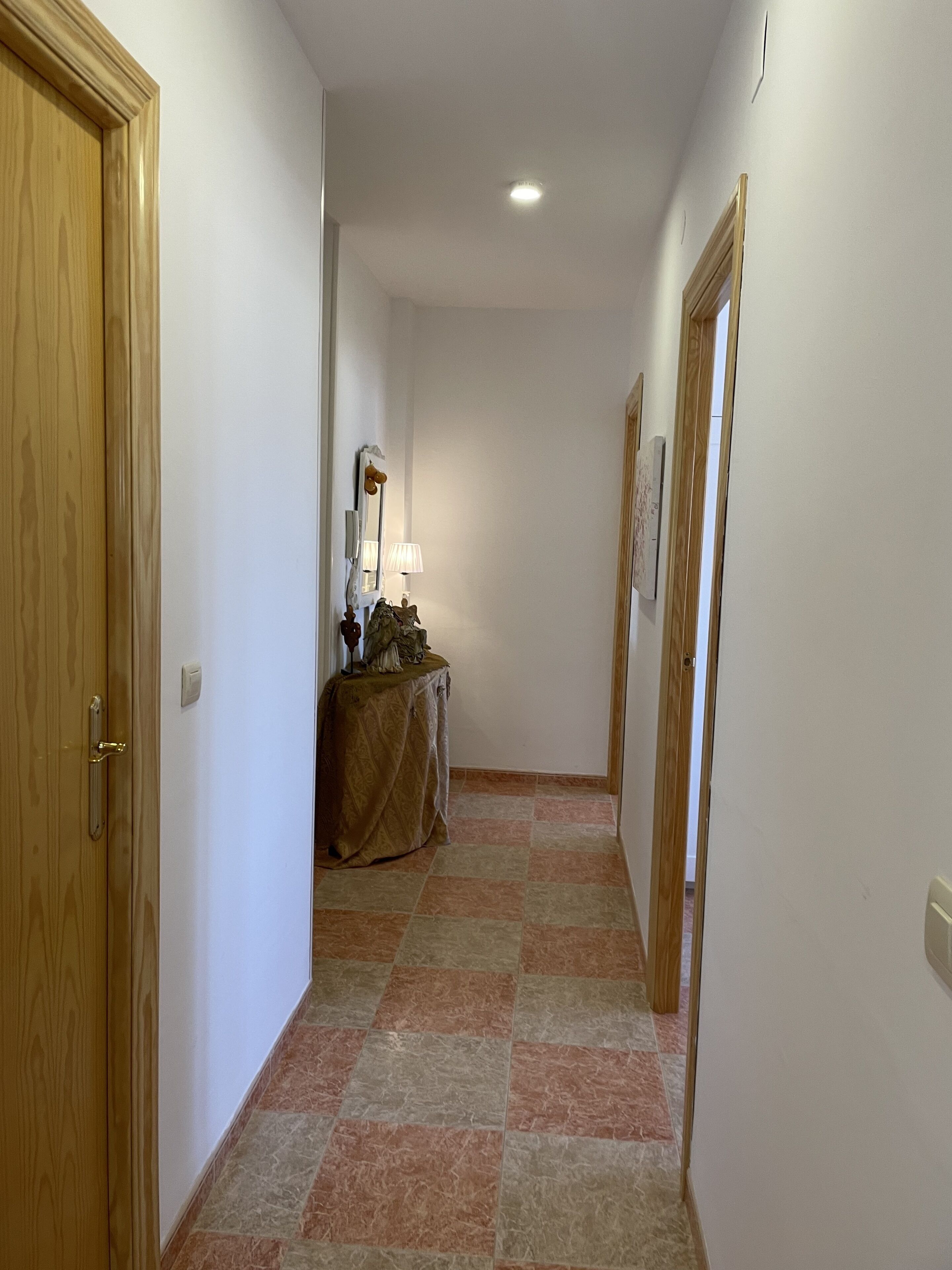 A narrow hallway with wooden doors on both sides, a tiled floor, and a small table with a lamp and decorative items in the center.