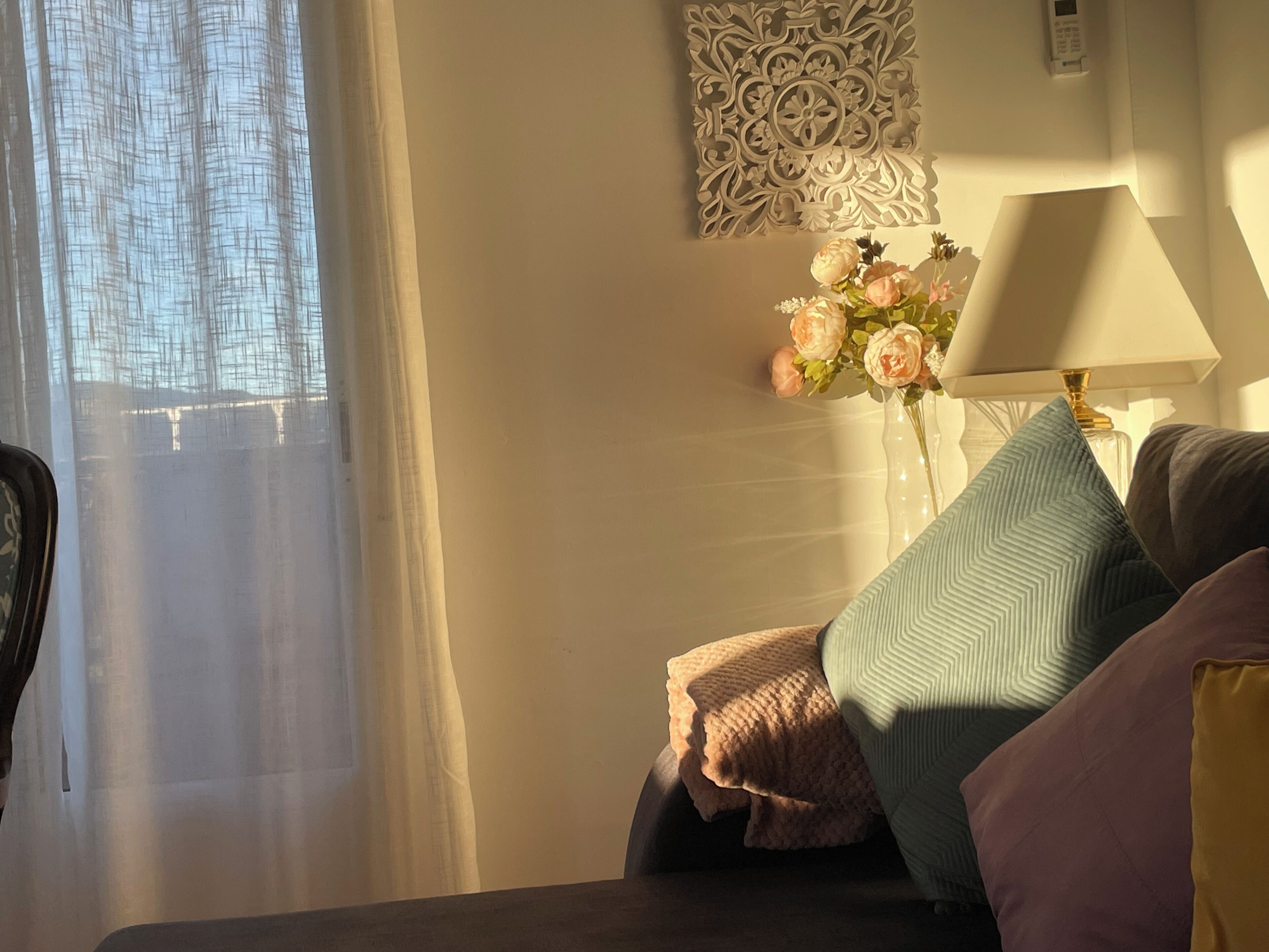 A bedroom with a bed, a bedside table with a lamp, a decorative wall piece, and a window with sheer curtains.