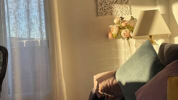 A bedroom with a bed, a bedside table with a lamp, a decorative wall piece, and a window with sheer curtains.