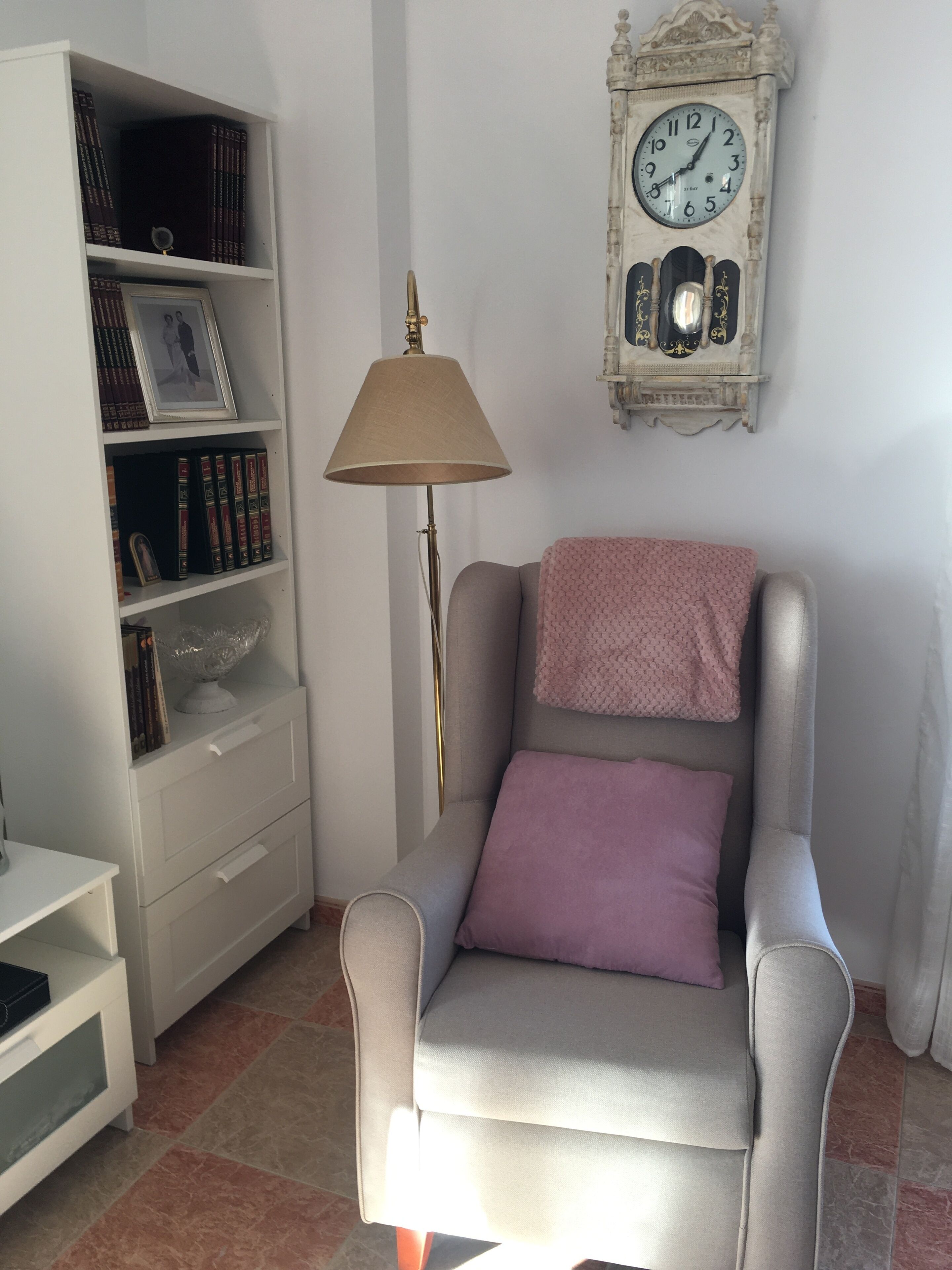 Un salon avec un fauteuil gris, une bibliothèque blanche, une lampe sur pied et une horloge murale.