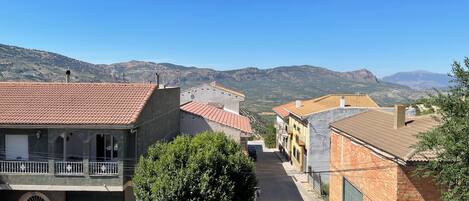 A narrow street with traditional buildings, a balcony with a railing, and a mountainous backdrop.