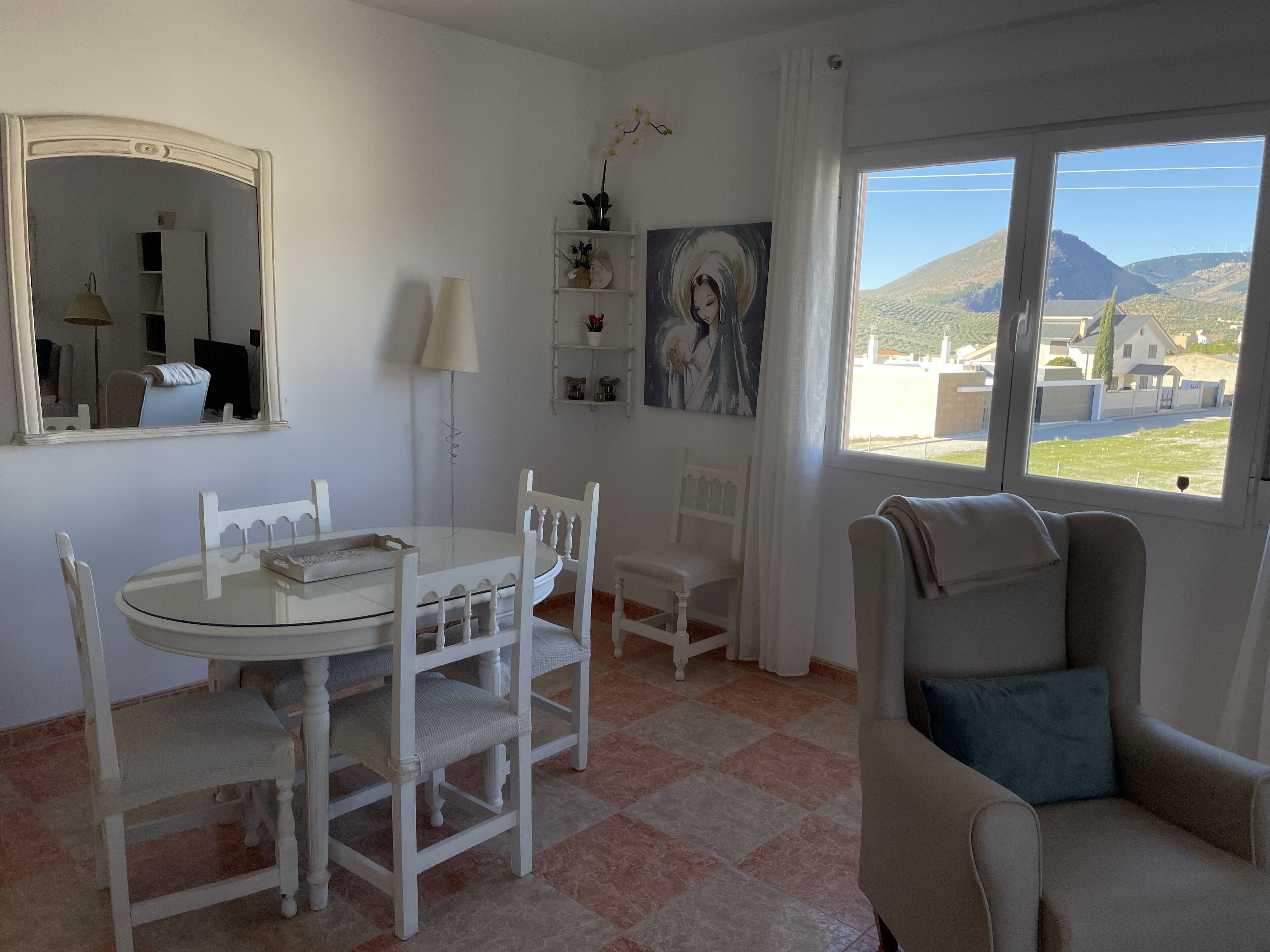 Une pièce avec une table ronde, des chaises blanches, un canapé, une grande fenêtre donnant sur des collines, un miroir et un tableau accroché au mur.