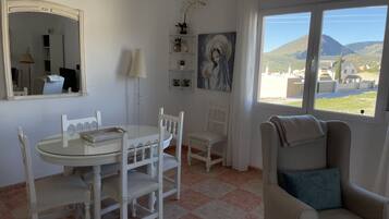 Une pièce avec une table ronde, des chaises blanches, un canapé, une grande fenêtre donnant sur des collines, un miroir et un tableau accroché au mur.