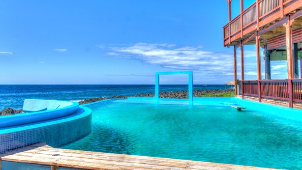 Una piscina cristallina blu con un pontile in legno, una struttura in legno con un balcone e vista sull'oceano.
