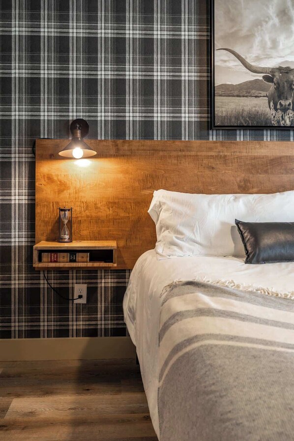 A bedroom with a wooden headboard, a bedside lamp, a white bed, a black pillow, and a framed picture of a bull.