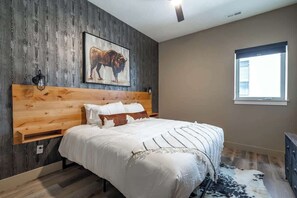 Ein Schlafzimmer mit einem Holz-Kopfteil, einem großen Bett mit weißer Bettwäsche, einem Teppich mit Kuhmuster, einem Fenster mit Jalousien und einer Wandleuchte.