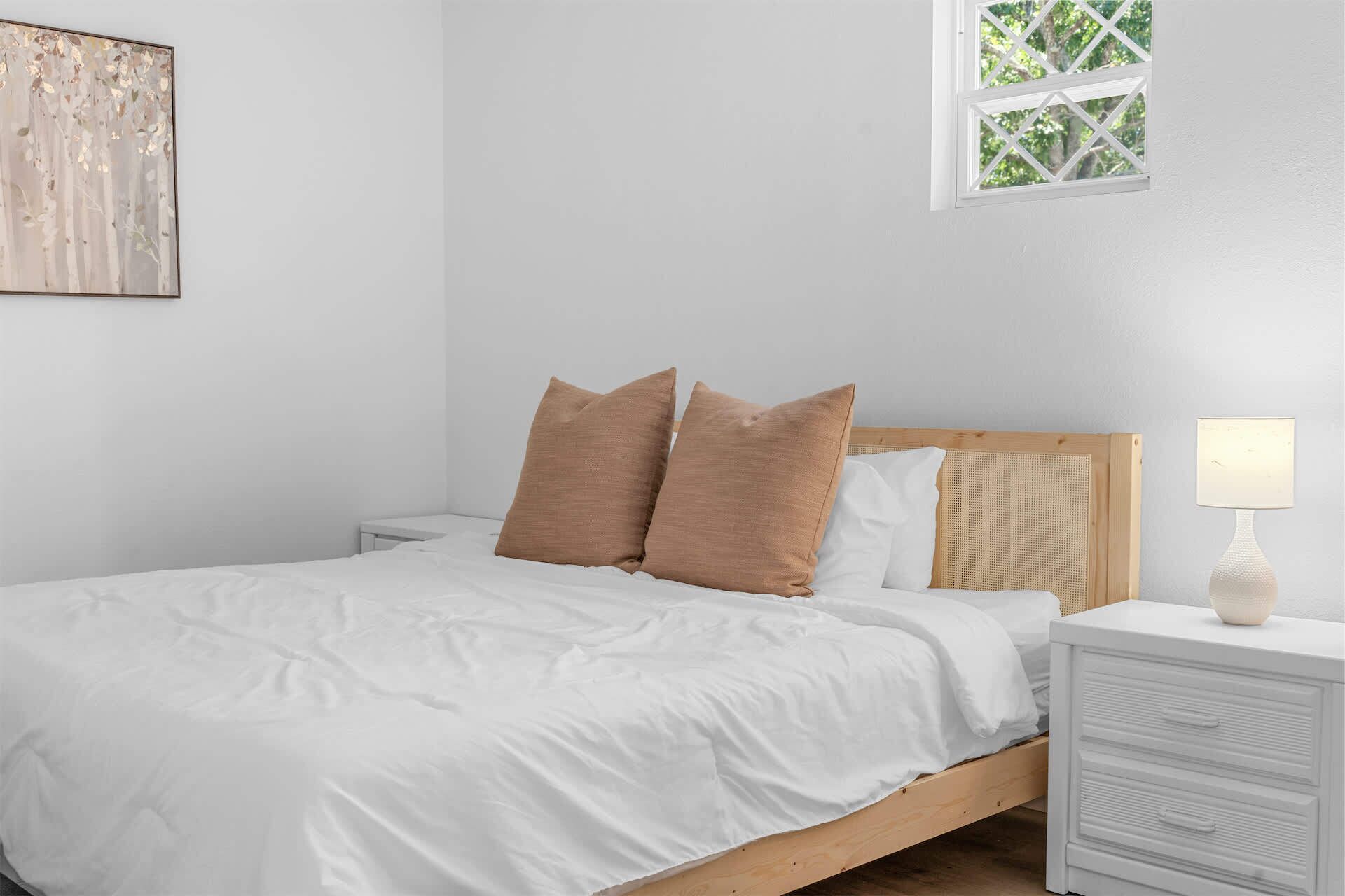 A bedroom with a bed, pillows, a nightstand, a lamp, a window, and a wall art piece.