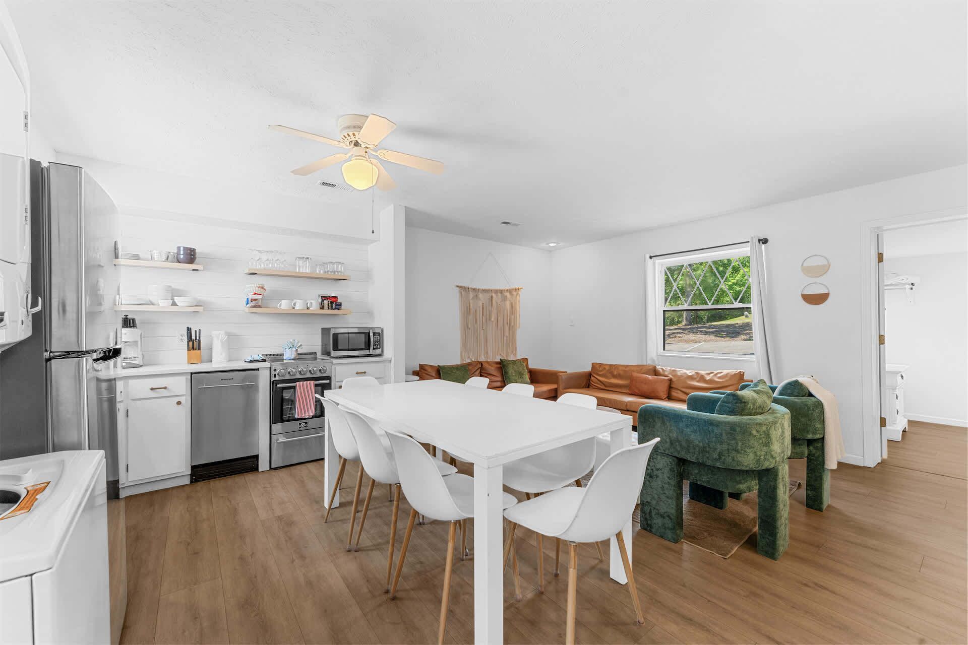 A modern kitchen with white cabinetry, a dining area with a white table and chairs, and a living room with a green sofa and a window with a view.