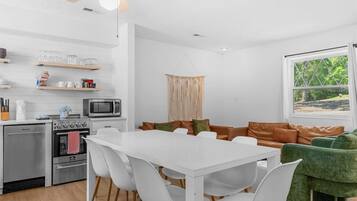 A modern kitchen with a microwave, oven, and coffee maker, a dining area with a white table and chairs, and a green sofa.