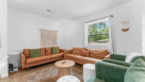 Un salon moderne avec un canapé en cuir, des fauteuils verts, une table basse ronde et une grande fenêtre donnant sur un paysage verdoyant.