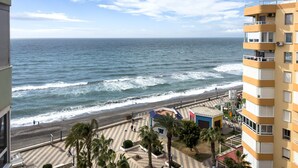 Vista costera con playa, un paseo marítimo con palmeras y un edificio de varios pisos con balcones.