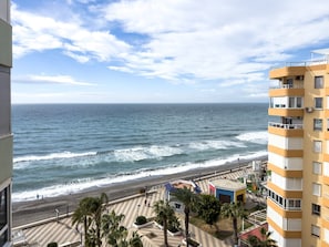 A coastal view with a beach, a promenade with palm trees, and a multi-story building with balconies.
