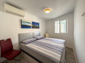 Una camera da letto con un letto, una sedia, un condizionatore, un quadro, una finestra e un lampadario.