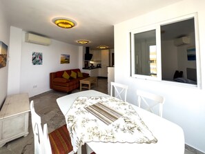 Un ambiente compatto con zona pranzo, divano e angolo cottura sullo sfondo, oltre a una finestra.