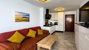 Un espacio habitable compacto con sofá, una mesita de madera, una zona de cocina y una puerta que da a otra habitación.