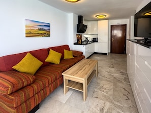 Un espace de vie compact comprenant un canapé, une petite table en bois, un coin cuisine et une porte donnant sur une autre pièce.