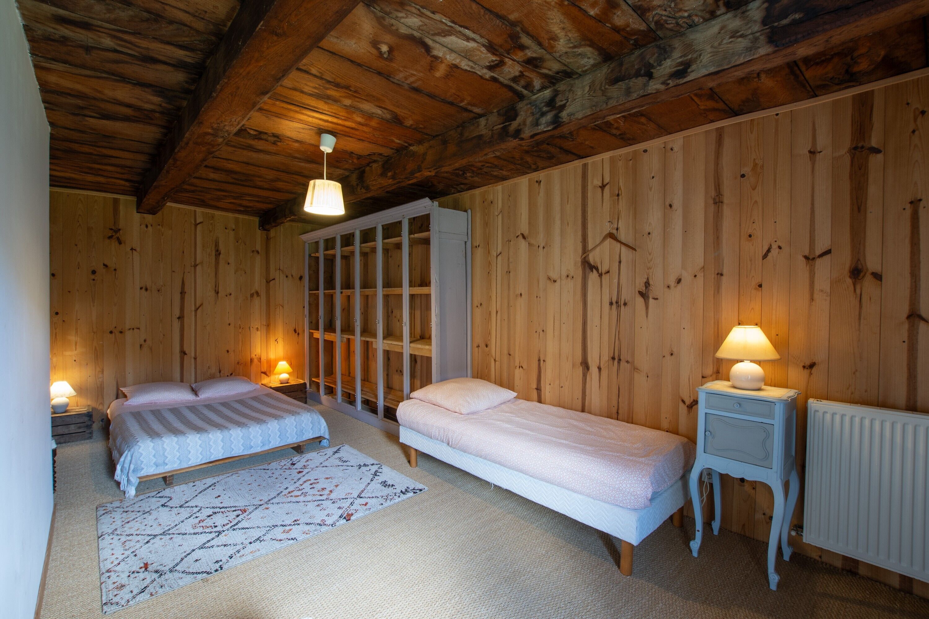A wooden-paneled room with two beds, a white wardrobe, and a small bedside table with a lamp.