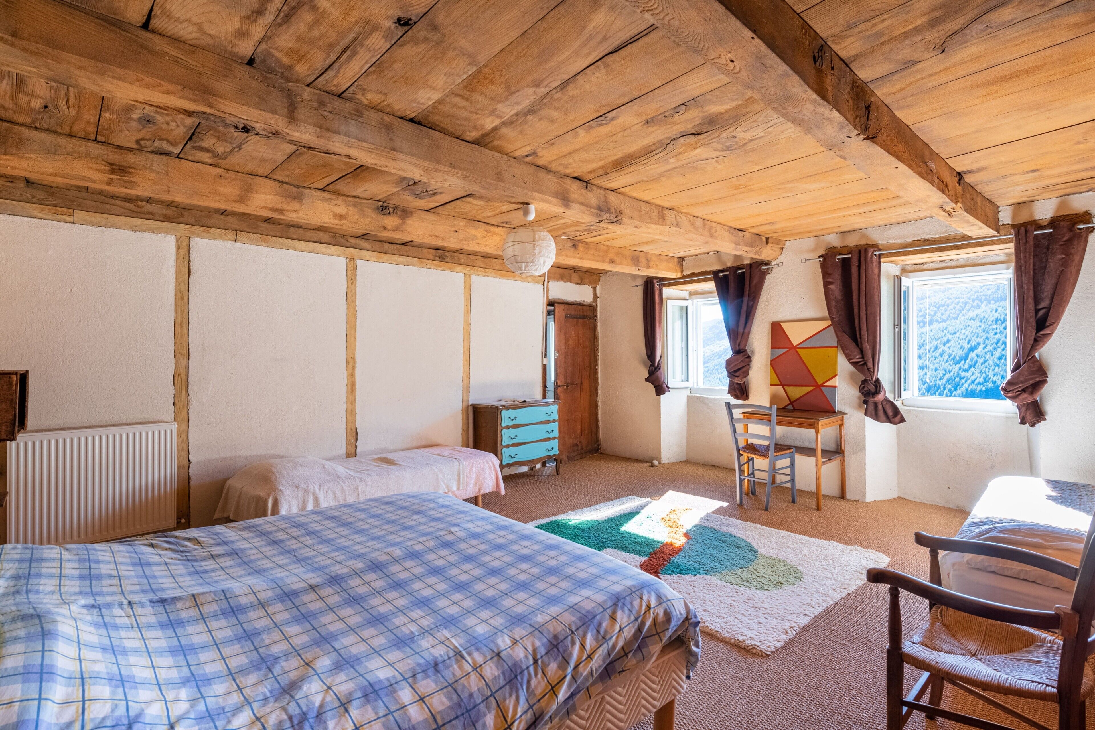 A room with wooden beams, a bed, a dresser, a chair, and a window with curtains.