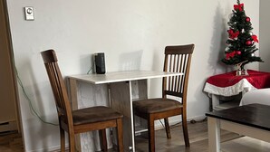 Un comedor con una mesa blanca y dos sillas de madera, una mesita con mantel rojo y blanco, y un árbol de Navidad adornado con adornos rojos y plateados.