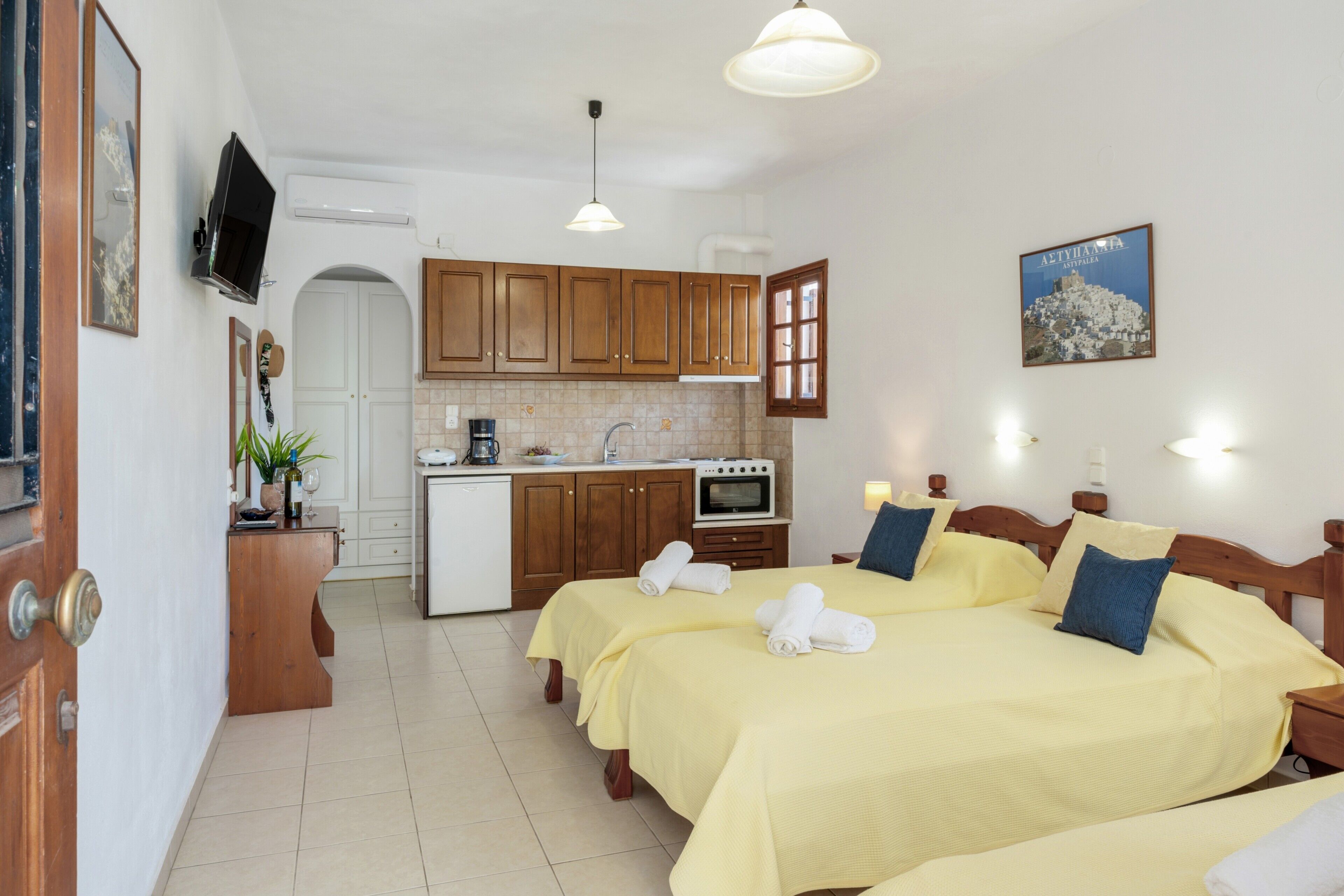 A hotel room with two beds, a kitchenette, a TV, and a painting on the wall.