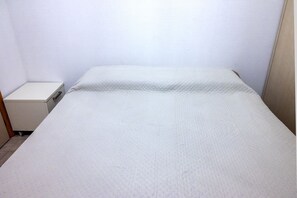 A small, single-bed room with a plain white wall and a simple bedside table.