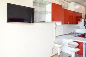A modern kitchen with a flat-screen TV, white and red cabinets, a sink, and two white bar stools.