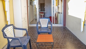 Un patio con dos sillas y una mesa pequeña, una puerta corrediza de vidrio que da a un espacio interior, y una pared amarilla a la izquierda.