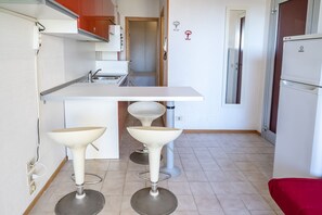 A compact kitchen with a white countertop, two bar stools, a sink, and a refrigerator.