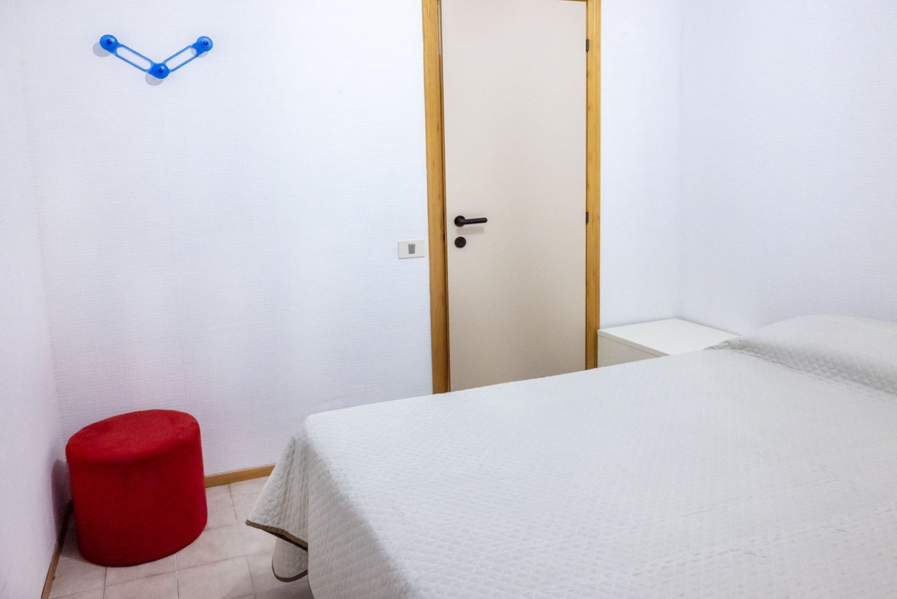Une pièce avec un lit, une porte, un tabouret rouge et un crochet fixé au mur.