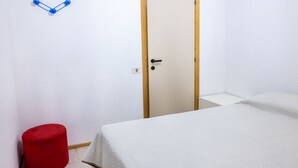 Une pièce avec un lit, une porte, un tabouret rouge et un crochet fixé au mur.