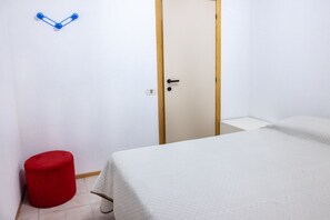 Um quarto com uma cama, uma porta, um banquinho vermelho e um gancho fixado na parede.