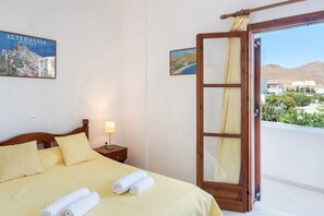 A hotel room with a bed, a nightstand, a lamp, a window with a view of a mountain and buildings, and a door leading to a balcony.