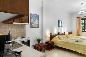 Una cocina con estufa, cafetera y una planta en maceta. Hay dos camas con ropa de cama amarilla y un ventanal con cortinas.