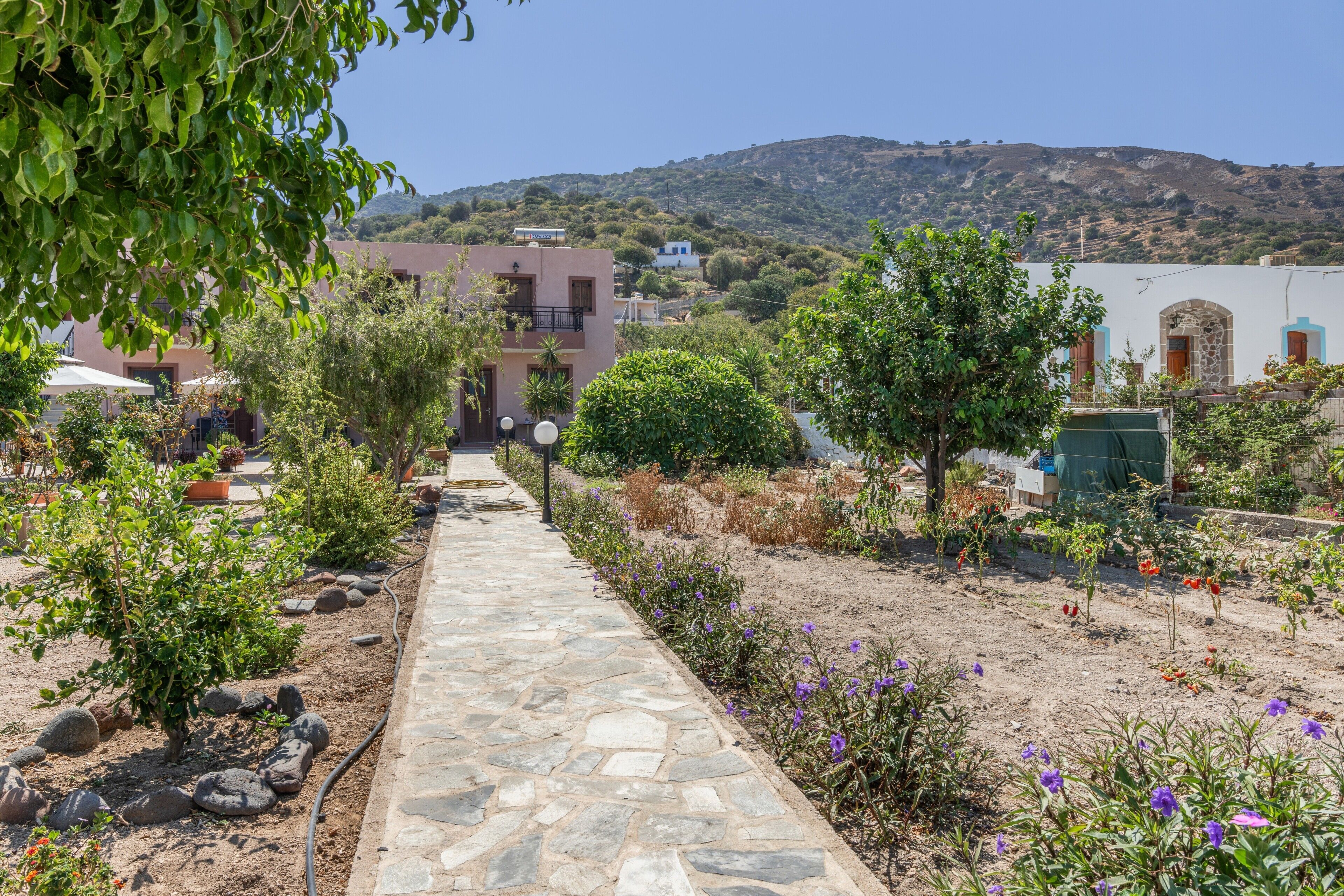 A stone pathway leading to a building with a balcony, surrounded by greenery and a hilly landscape.
