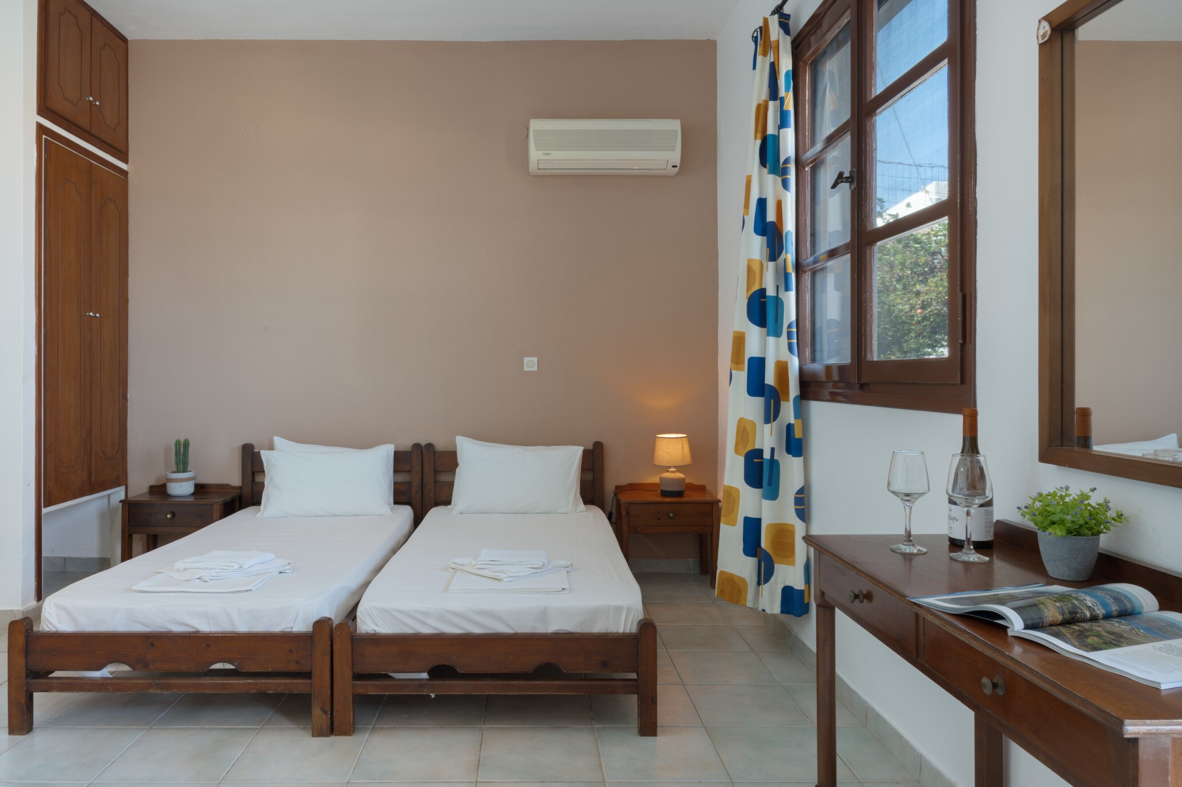 Ein Schlafzimmer mit zwei Einzelbetten, einem Holztisch, einem Spiegel, einem Fenster mit Vorhängen, einer Klimaanlage und einem Nachttisch mit einer Lampe.
