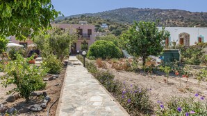 A stone pathway leading to a building with a balcony, surrounded by greenery and a hilly landscape.