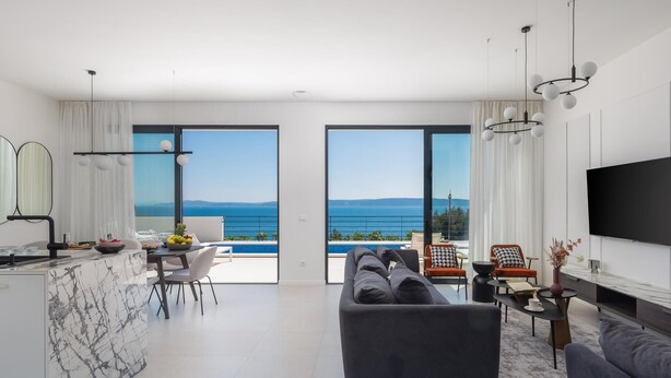Ein modernes Wohnzimmer mit einer großen Couch, einem Essbereich mit Tisch und Stühlen, einer Küche mit Marmorarbeitsplatte und einem großen Fernseher. Große Fenster bieten Blick aufs Meer.