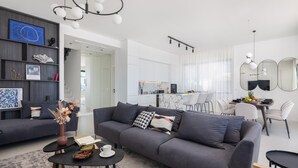 Ein modernes Wohnzimmer mit einem grauen Sofa, einem Couchtisch mit Büchern und einer Schale Obst, sowie einem Essbereich mit weißem Tisch und Stühlen.