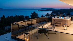 Eine moderne Außenterrasse mit Glastrennwand, Sitzgelegenheiten und einem Whirlpool mit Blick auf eine Küstenlandschaft im Abendrot.