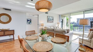 Un salon moderne avec une table à manger en verre, des chaises en osier, un canapé et une grande fenêtre donnant sur l’extérieur.