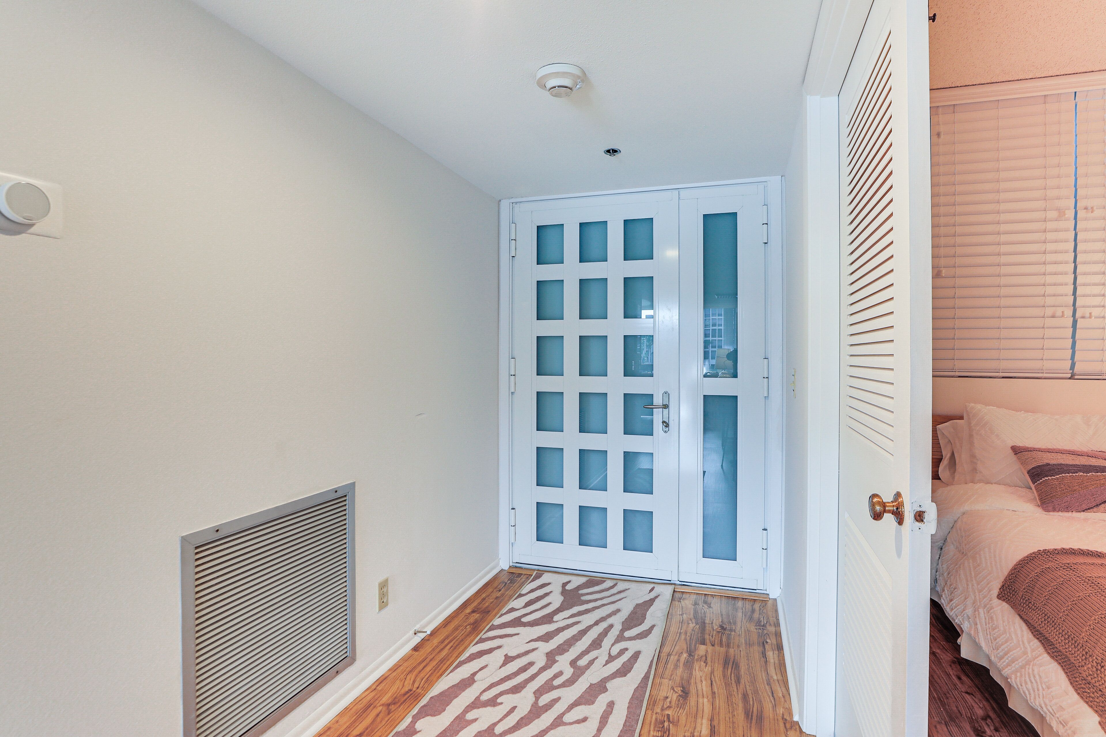 A narrow hallway with a white door, a vent on the left, and a bed with a patterned bedspread on the right.