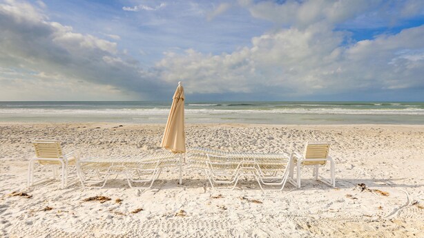 Une plage avec des chaises longues blanches et un parasol beige, donnant sur l’océan.
