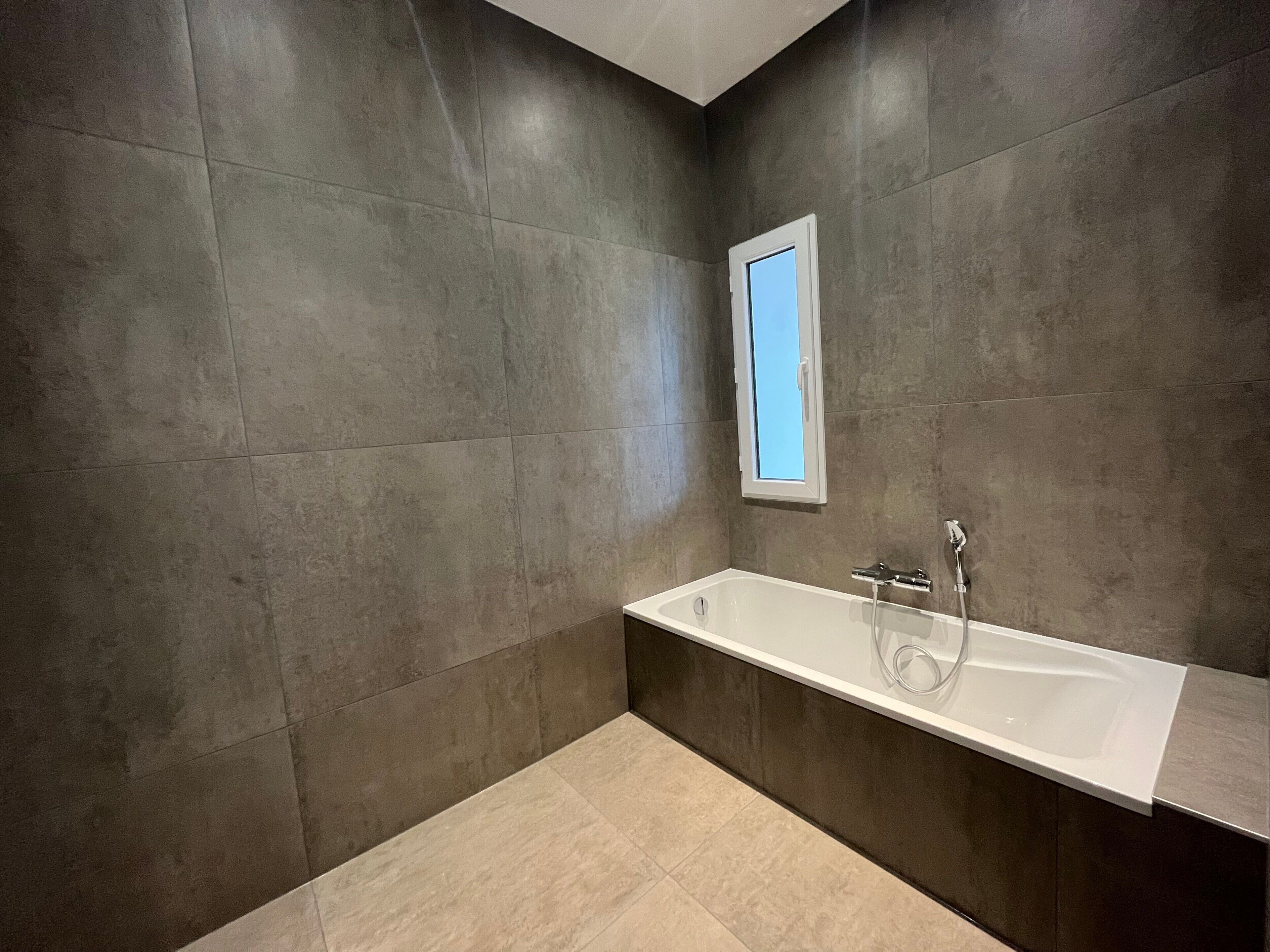 A modern bathroom with a large bathtub, a window, and dark tiled walls.