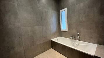 A modern bathroom with a large bathtub, a window, and dark tiled walls.