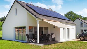 Una casa moderna con paneles solares en el techo, un porche cubierto con mobiliario exterior y un coche estacionado cerca.