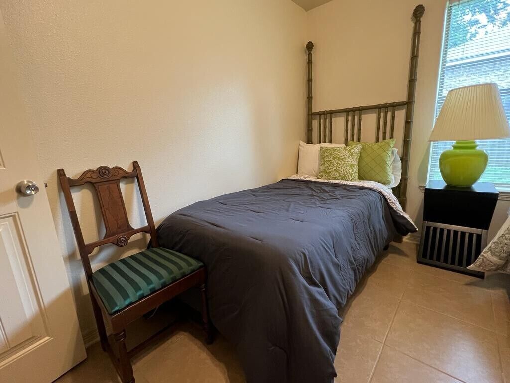 A bedroom with a bed, a chair, a nightstand with a lamp, and a window with blinds.