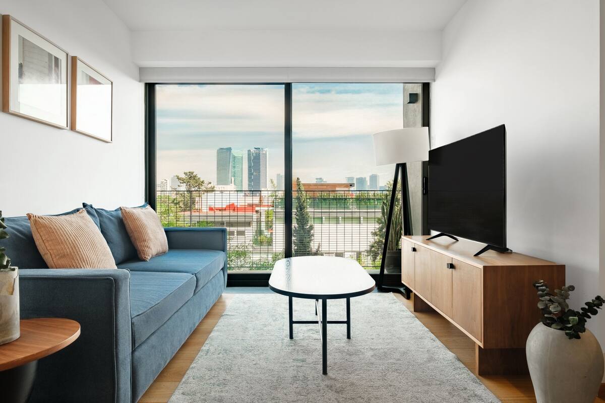A modern living room with a sofa, a small table, a TV on a wooden cabinet, and a large window with a city view.